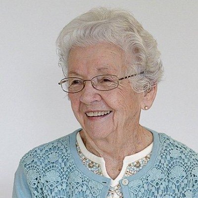 older woman with grey hair, blue cardigan, and glasses is smiling in a portrait shot